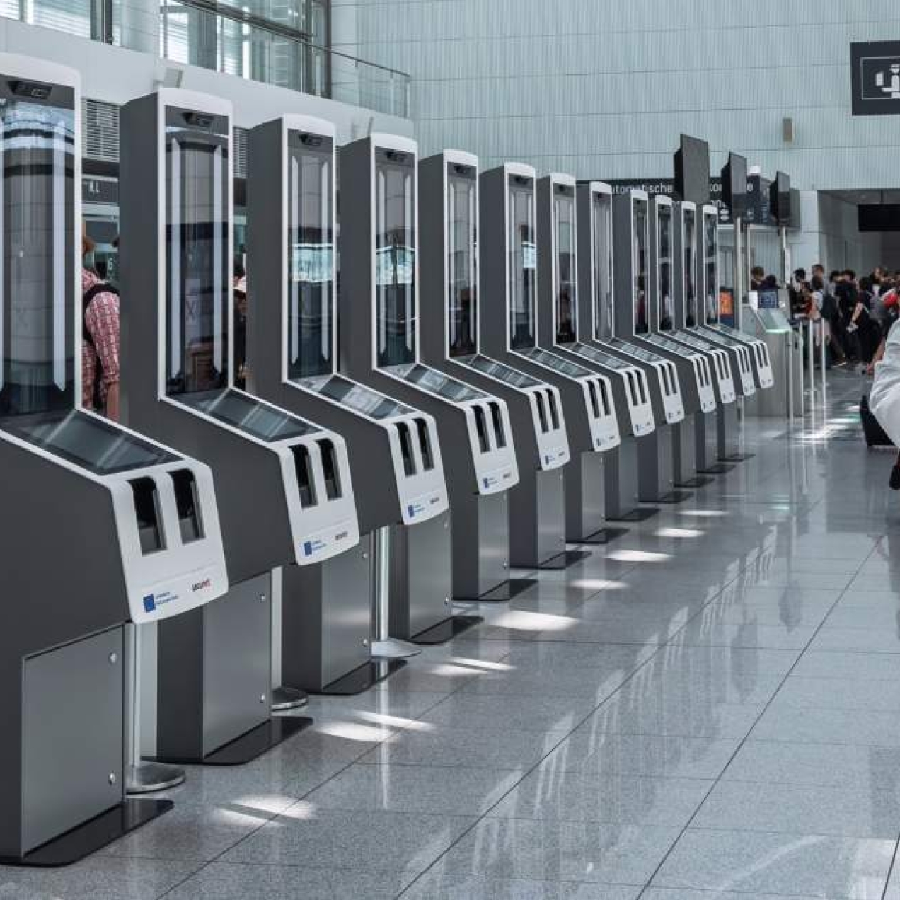 The back of a person wearing a blue top and white trousers looking at a row of tall, grey coloured computer scanners in a spacious airport.