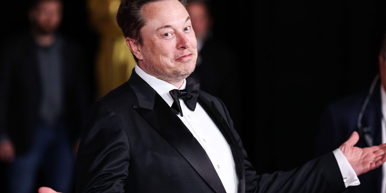 Elon Musk, a white man with short brown hair, looks at the camera with his arms open, while wearing a black suit and bow tie. The background is mostly dark with glimpsed of people behind him