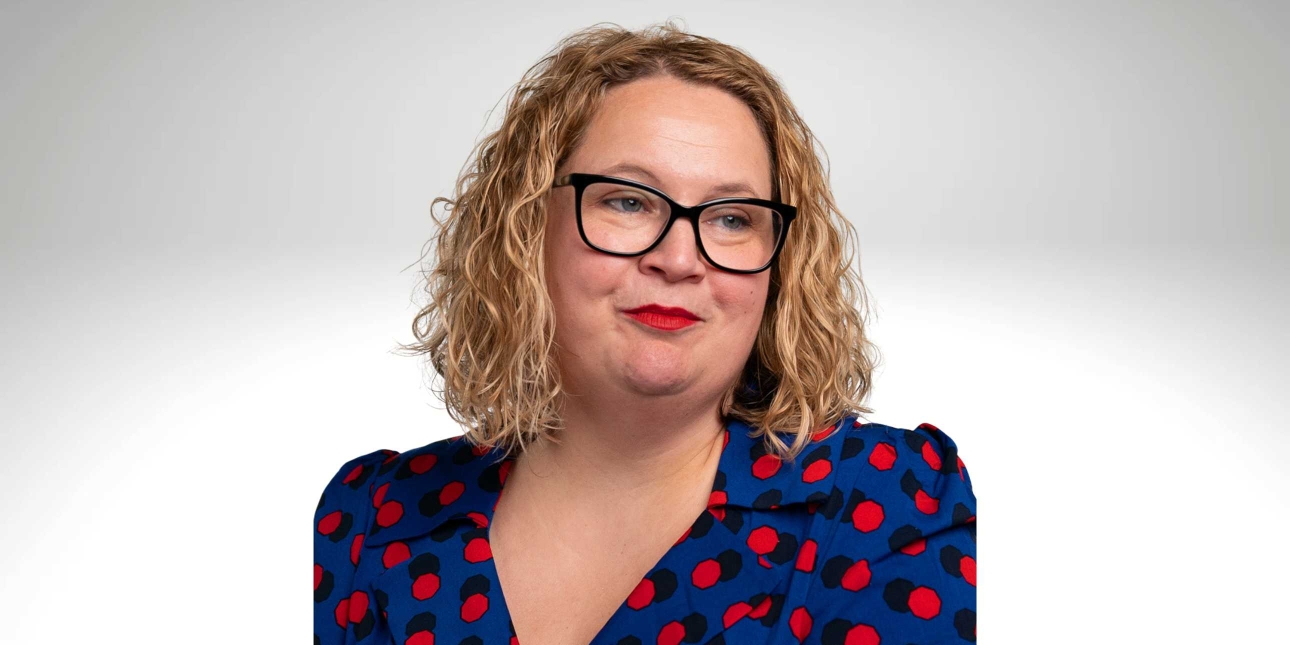 A side portrait of Emma Duke, a white woman with blonde shoulder length hair, who wears glasses and a red and black polka dot blue top
