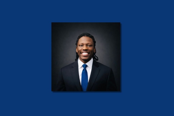 A colour portrait on a blue background of Emmanuel Kisolom smiling. Emmanuel is a Black man who is wearing a dark suit and a navy tie.
