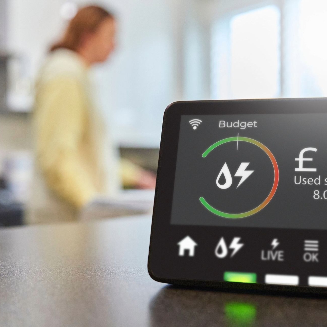 A close up image of a smart meter device showing energy consumption for the day. In the background is a blurred image of a woman in a kitchen