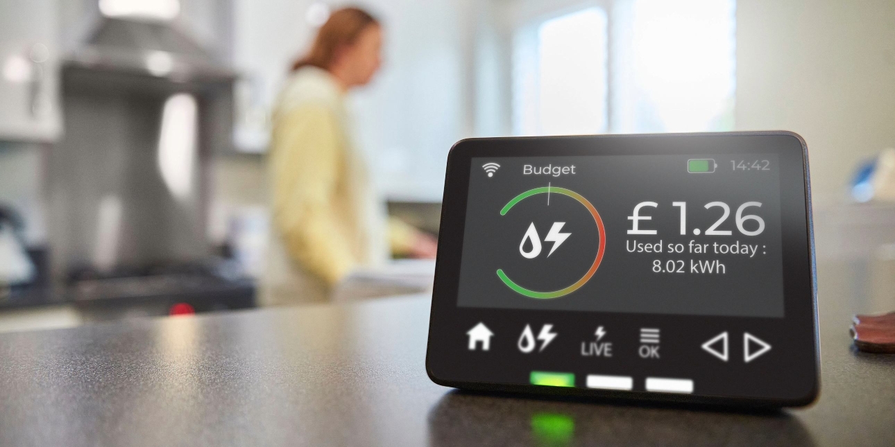 A close up image of a smart meter device showing energy consumption for the day. In the background is a blurred image of a woman in a kitchen