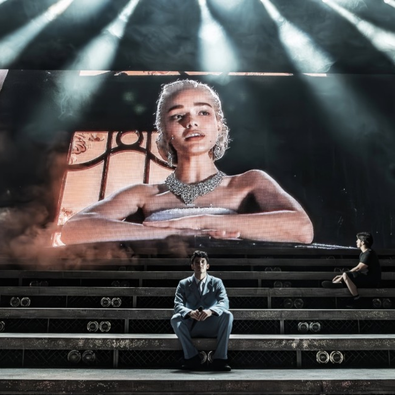 Rachel Zegler leans over a balcony on a large screen at the back of a stage. In front of her on steps sit two men.