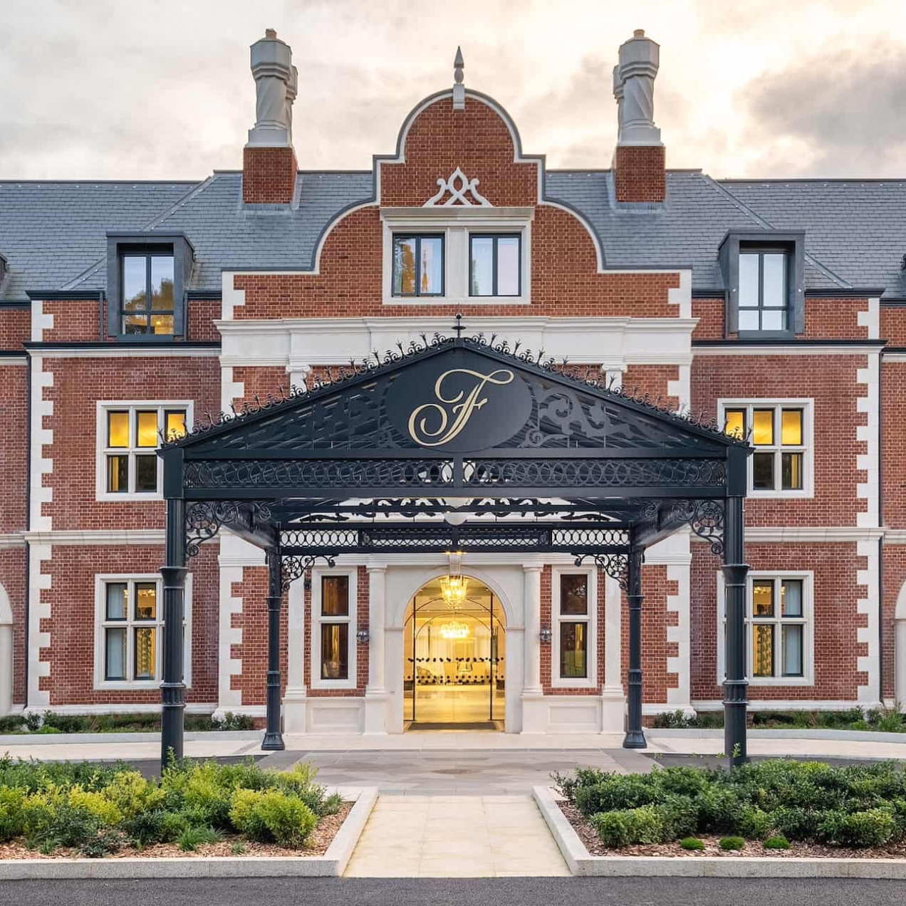 The exterior entrance of Fairmont Windsor Park, an ornate three storey red brick building with a dozen windows, eight chimneys and a decorate black canopy embossed with an italic F covering a portion of the green vehicle turning circle out front. It