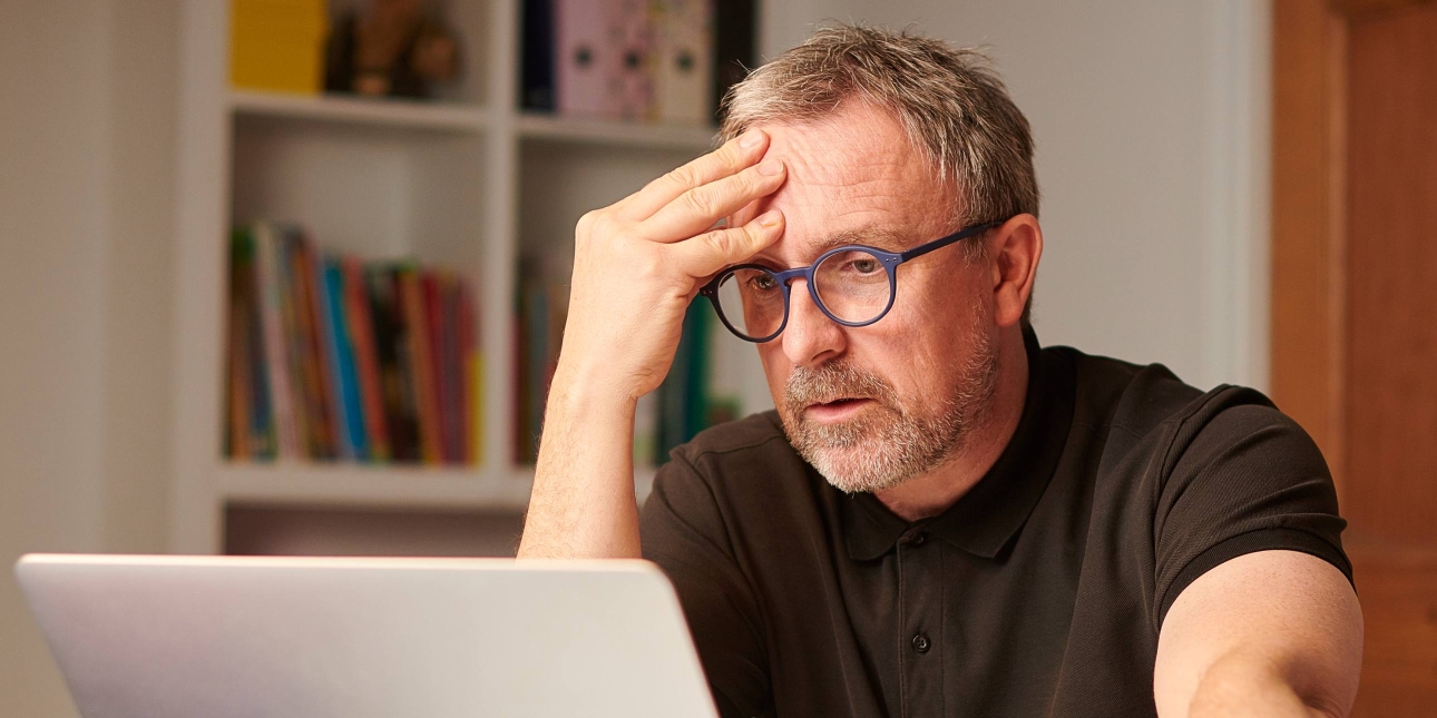 An anxuous white middle-aged man with short hair and glasses hold his forehead while sat in front of a laptop