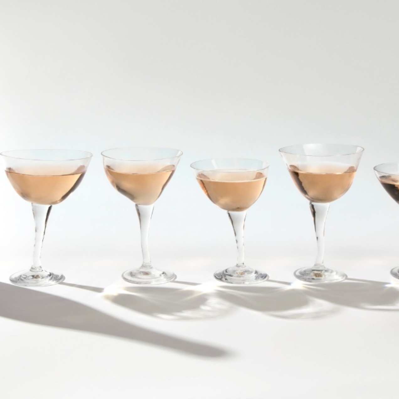 A group shot on a white background of a dark uncorked Folc wine bottle alongside five different-shaped stem glasses of sparkling rosé wine