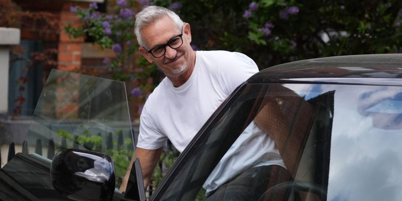 Gary Lineker, dressed in a white t-shirt, getting into the driver's seat of his black car. Lineker is a white man with grey hair and glasses. There are red brick houses in the background.