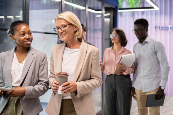 Walking through an office corridor, a young black business woman holding a book in deep conversation with an older white business woman holding a coffee cup.