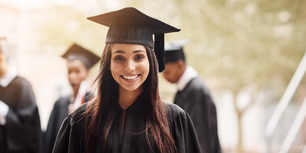 Portrait of a young smiling Asian woman wearing a black graduation gown and mortarboard hat. Out of focus in the background are three fellow graduates