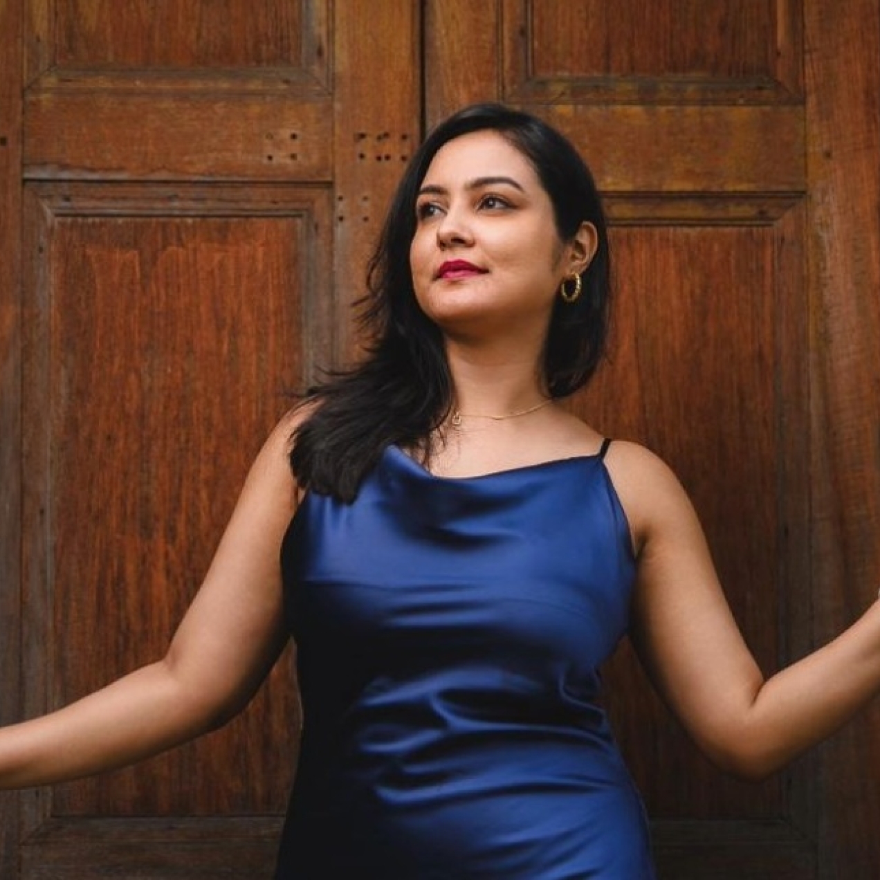 Hilla Lakhpatwala wearing a blue dress in front of wooden door with her arms reaching out to the stone porch. Hilla is an Asian woman with long dark hair.