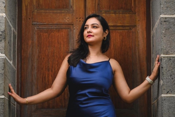 Hilla Lakhpatwala wearing a blue dress in front of wooden door with her arms reaching out to the stone porch. Hilla is an Asian woman with long dark hair.