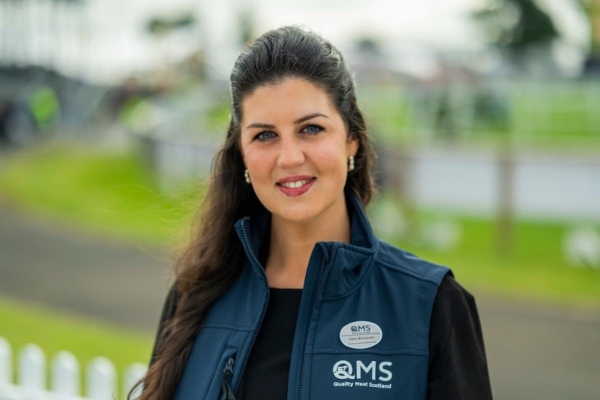 A colour portrait of Holly McLennan. Holly is a white woman with long dark hair wearing a blue gilet with writing on it. There is greenery behind her.