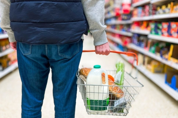 The back of a man in a supermarket aisle wearing a blue gilet and blue jeans holding a shopping basket full of bread, milk and groceries.