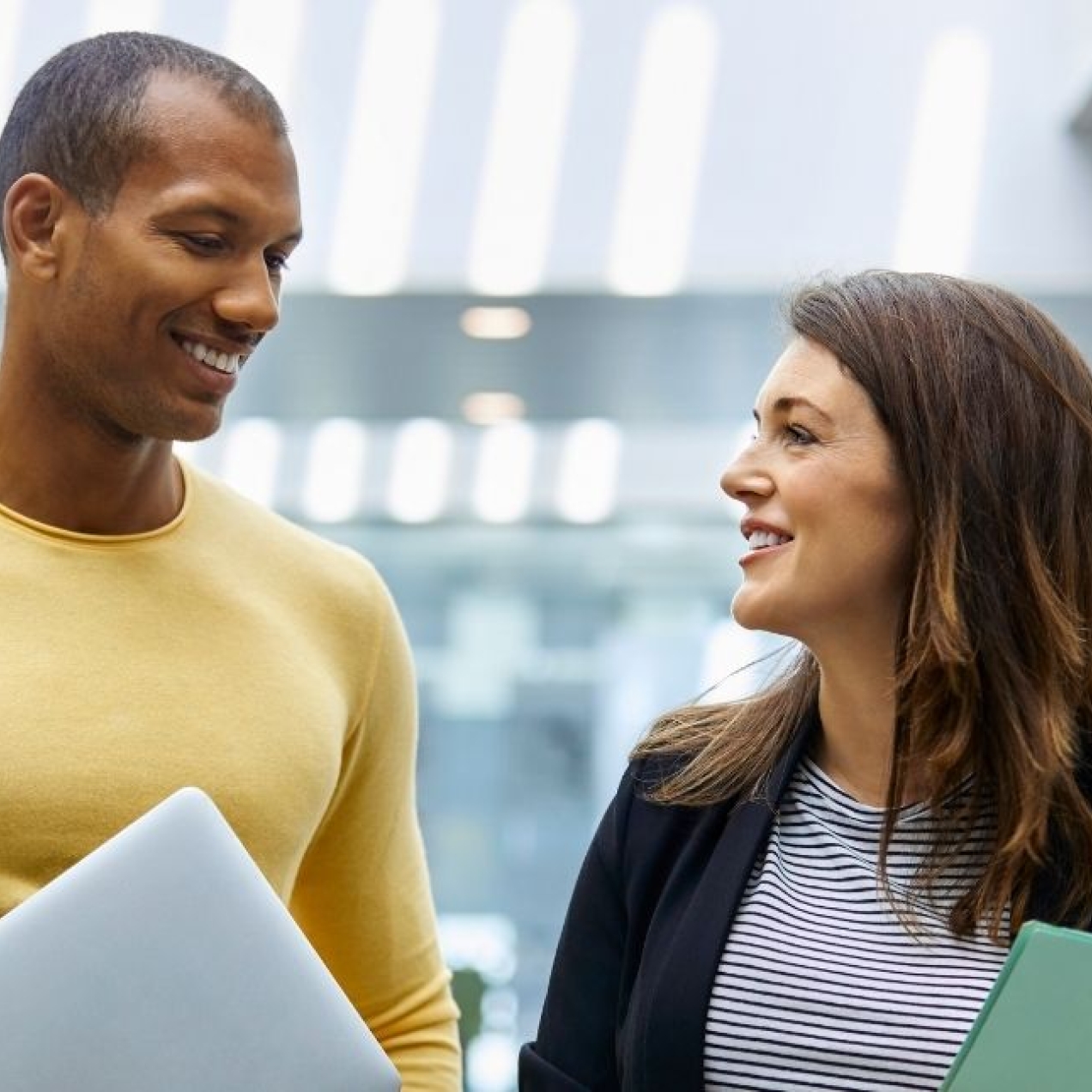 Two smiling colleagues - a Black man wearing a yellow jumper and a white woman wearing a dark jacket - chat in a light filled atrium. Both hold folders.