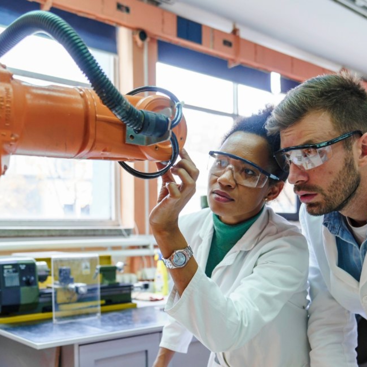 Two colleagues in white lab coats and protective glasses - a Black woman on the left and a white man on the right - inspect an orange robotic arm in a laboratory.