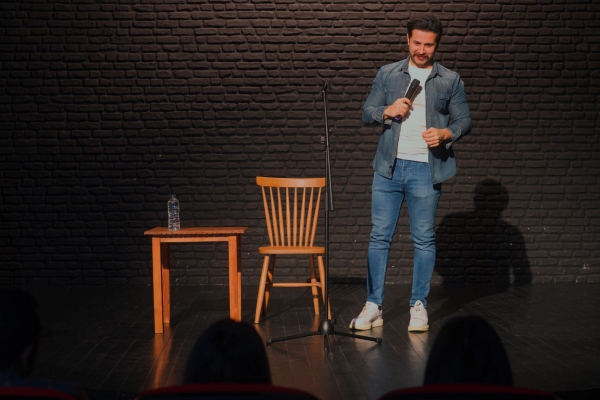 A white man with short dark hair and beard is stood on a stage holding a microphone facing a silhouetted audience