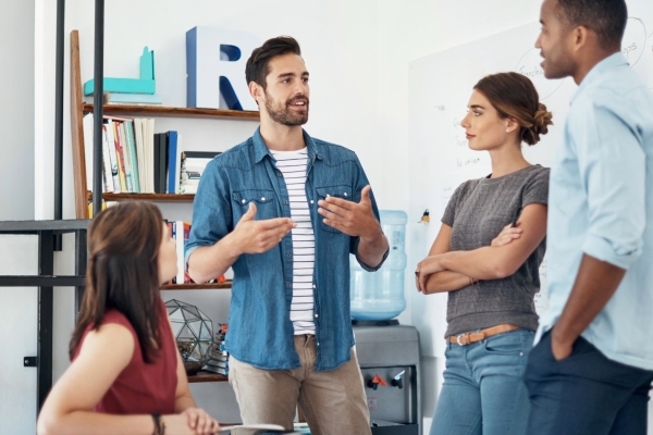 In a modern office, a white man with short dark hair and blue shirt is in discussion with three colleagues: a white woman and Black man who are stood up, and a white woman who is sat at a desk