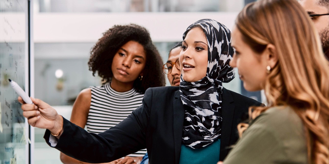 A woman wearing a black and white patterned hijab and black jacket writes on a board as colleagues of different genders and ethnicities look on