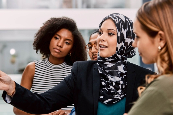 A woman wearing a black and white patterned hijab and black jacket writes on a board as colleagues of different genders and ethnicities look on