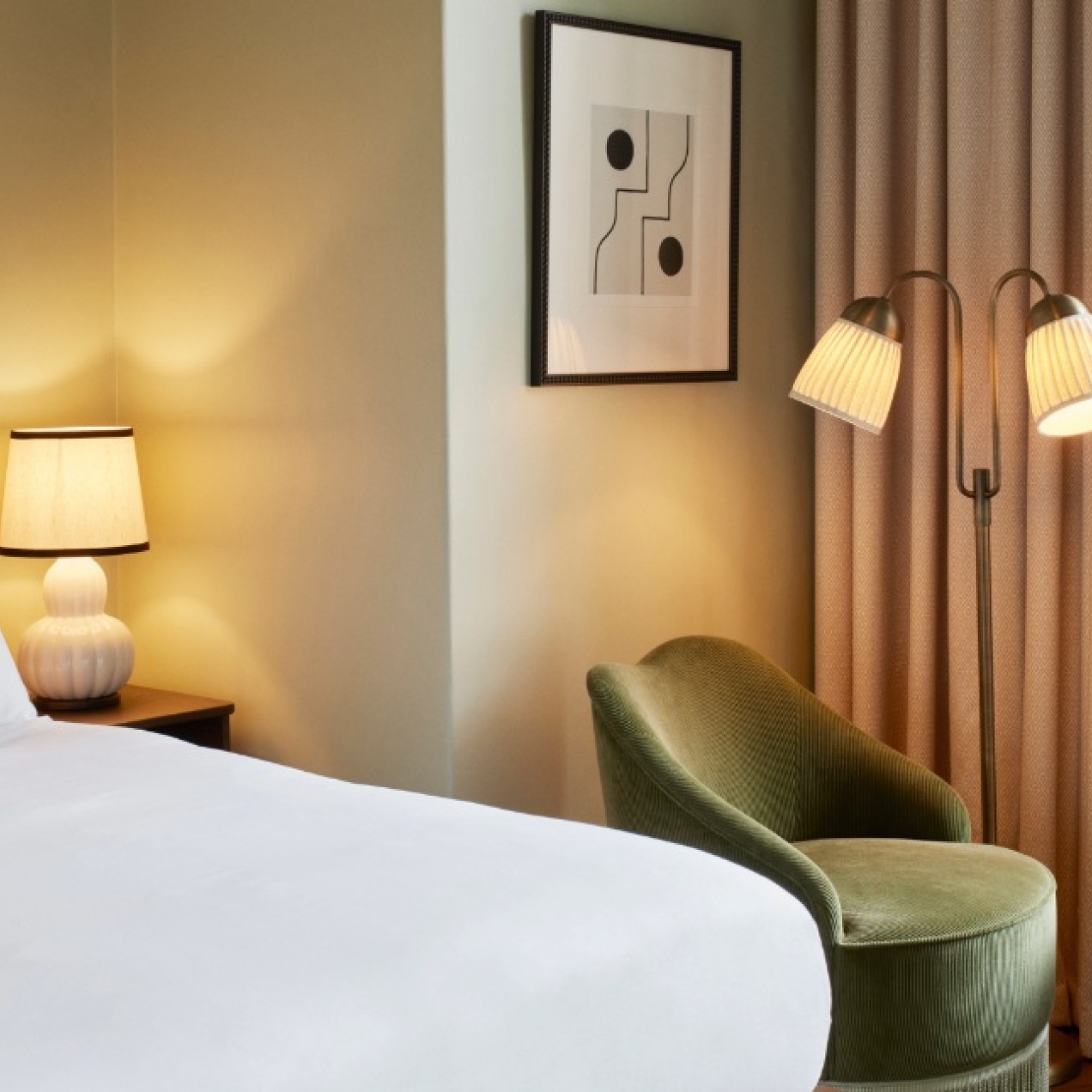 A bedroom featuring a large bed with white bedding and a red shell shaped headboard, plus a green high backed soft chair under a lamp stand in front of a window.