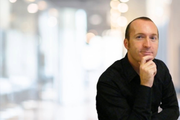 Against the blurred background of an office, Iain Patton sits with his right hand on his chin. Iain is a white man with receding dark hair who wears a black shirt. He is pictured off centre to the right of the image.