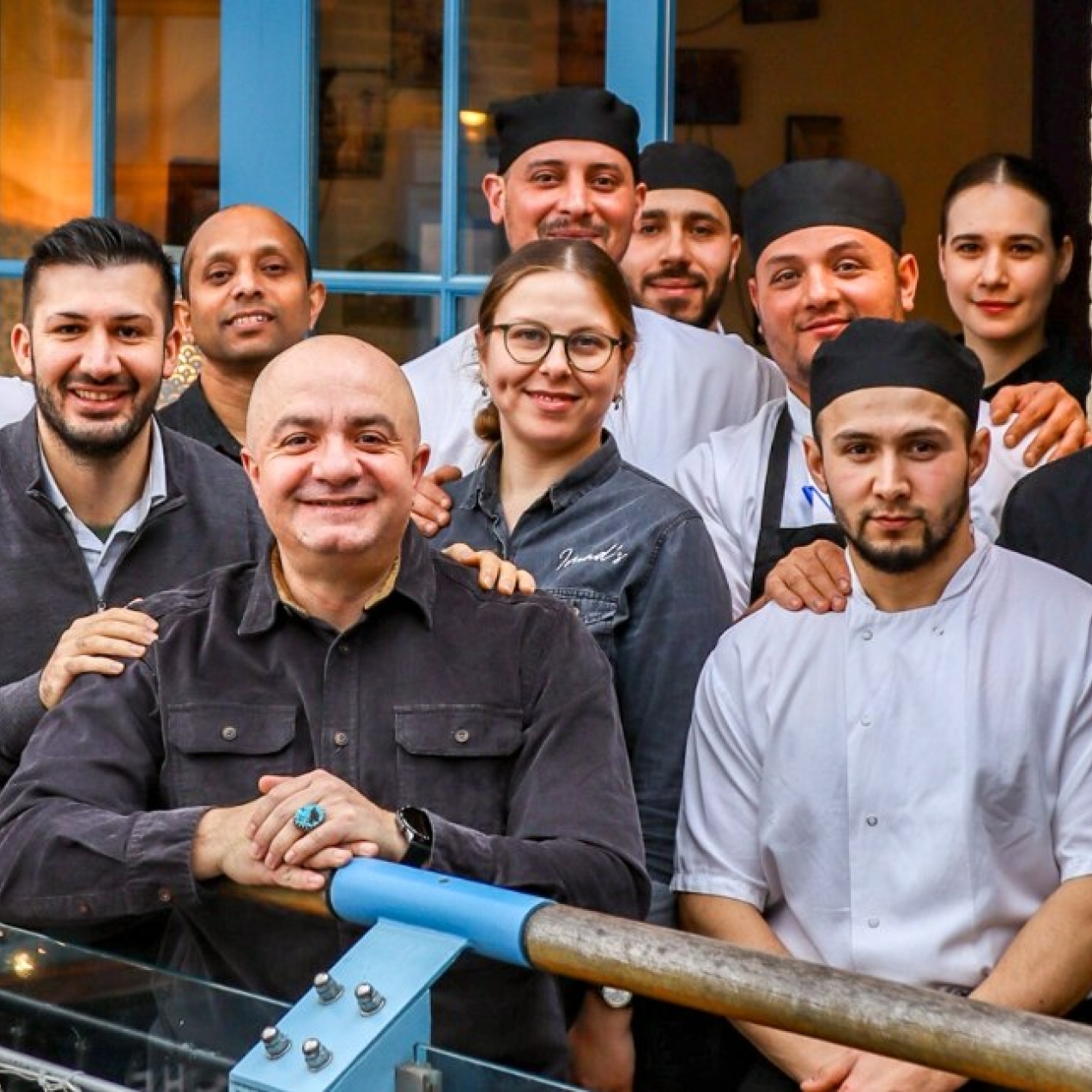 Thirteen people stood looking at the camera on a balcony outside a blue framed door. They mostly wear chefs whites or dark waiters' uniforms.