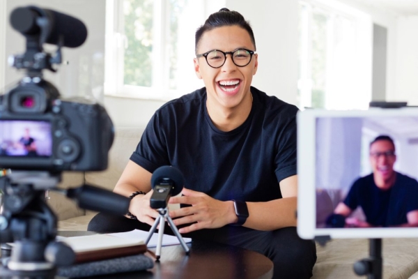 A smiling Asian man with short dark hair and dark shirt sits on a sofa in a modern home looking towards a camera and phone which are held on tripods..