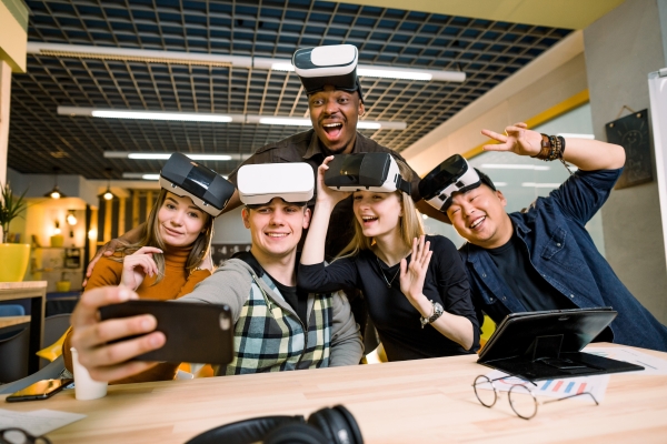 Five young colleagues in a modern office smile and gesture at a phone camera while wearing virtual reality headsets on their foreheads. The colleagues are two white women, one Asian man, one Black man and one white man.