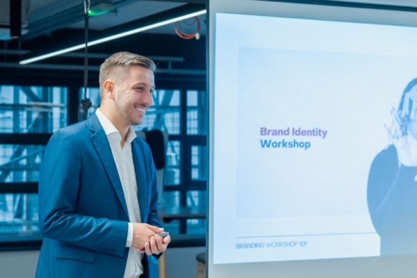 A colour portrait of a smiling Ivan Biošić stood side on to a display board. Ivan is a white man with short blond hair who is wearing a navy suit.