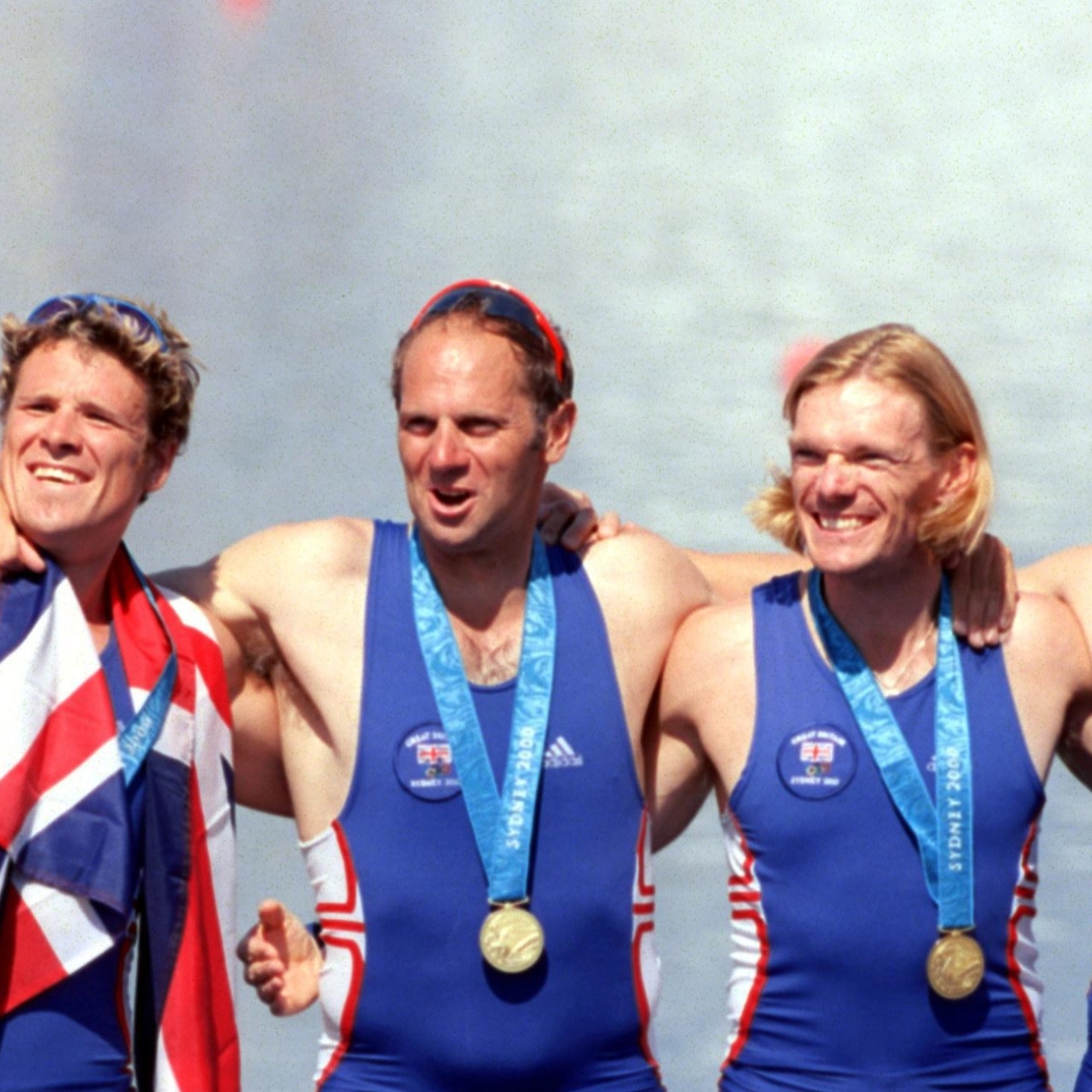 James Cracknell and team mates in a line with their arms around each other wearing blue swimsuits and displaying their gold medals with two of them punching their fists in the air