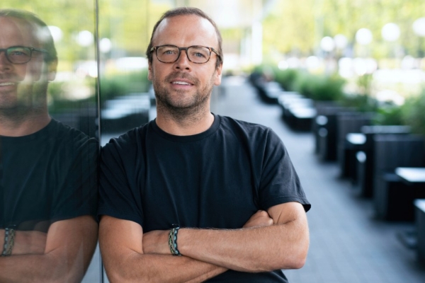 A colour portrait of James Peach leaning against a large glass window which shows his reflection. James is a white man with short dark hair and beard, wearing glasses and a black top.