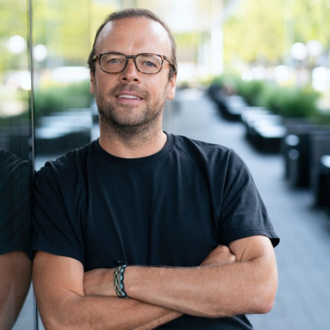 A colour portrait of James Peach leaning against a large glass window which shows his reflection. James is a white man with short dark hair and beard, wearing glasses and a black top.