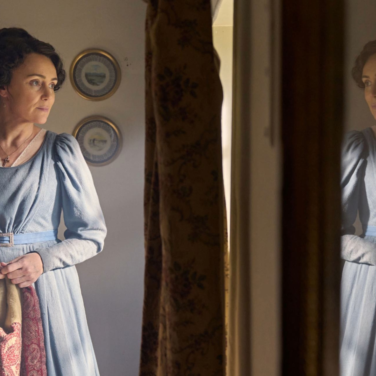 An episodic still from Miss Austen, featuring Keeley Hawes as Cassandra wearing a blue dress looking out of a bedroom window. Her reflection is on the right.
