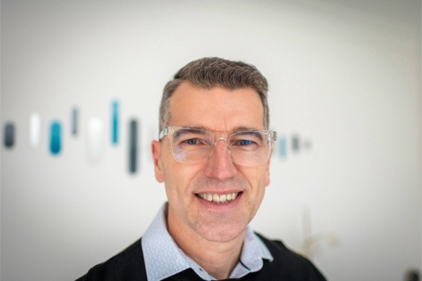 A portrait of Jon Large in front of a grey wall featuring blue-turquoise specks. Jon is a white man with short brown-grey hair, who wears transparent framed spectacles and blue collared shirt under a dark jumper