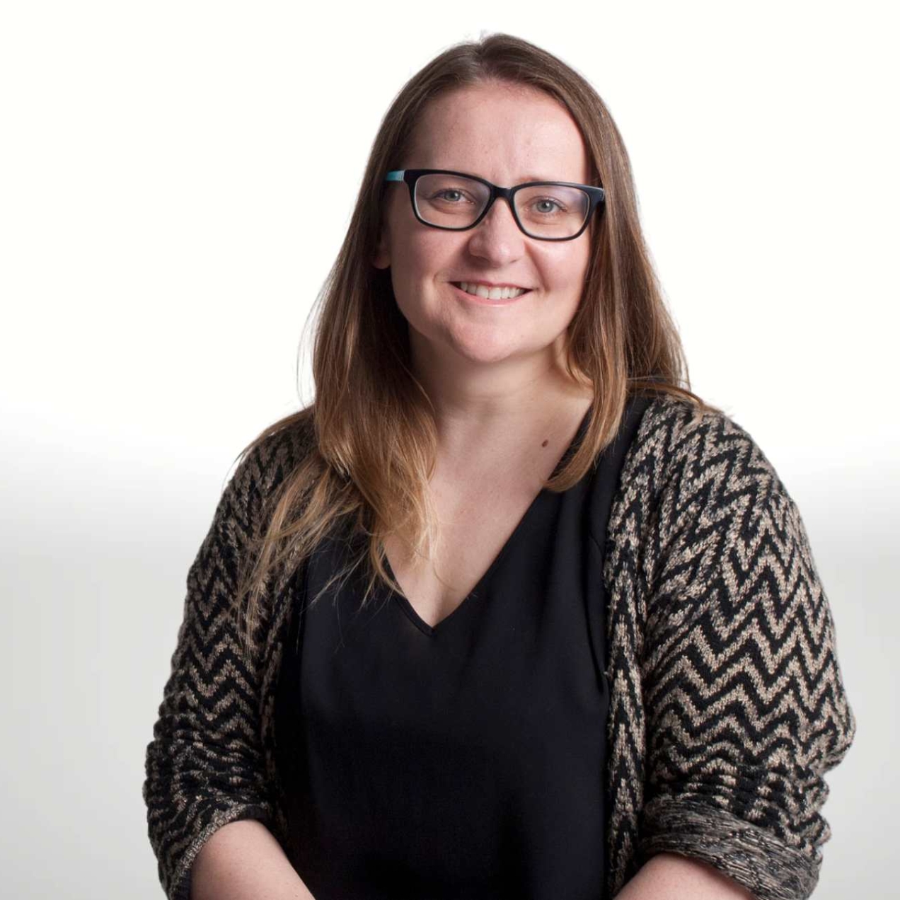 A portrait of a smiling Jule Wilson on a grey background. Jule is a white woman with long brown hair, glasses and wears a black and grey jacket over a black top