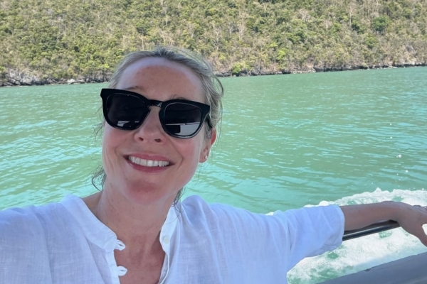 Juliet Kinsman is a blonde white woman wearing dark glasses.  She wears a white shirt and is sat with her hand over a rail with an expanse of water and the greenery of the shore behind her.