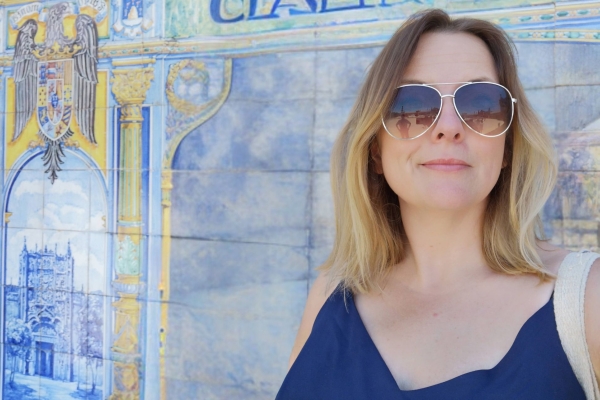 A close-up portrait of Kat Dibbits, a white woman with blonde hair and wearing glasses, against a blue and yellow tiled wall