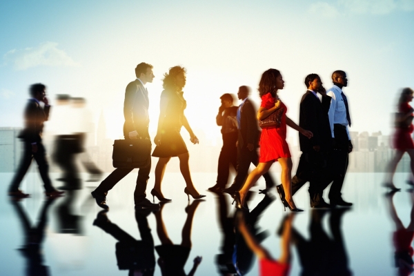 A variety of business people of different genders, ages and ethnicities walking along a reflective surface with the sun and skyscrapers behind them