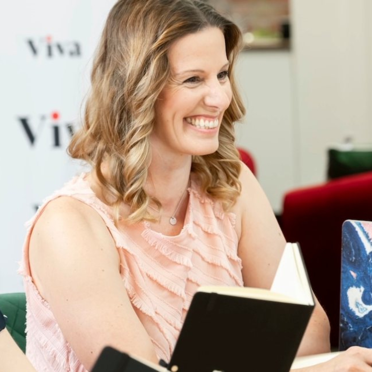 A smiling Lisa Egan-Woodhouse sat among members of the Viva team. Egan-Woodhouse is a white woman with shoulder length light brown hair, who is wearing a pink sleeveless top.