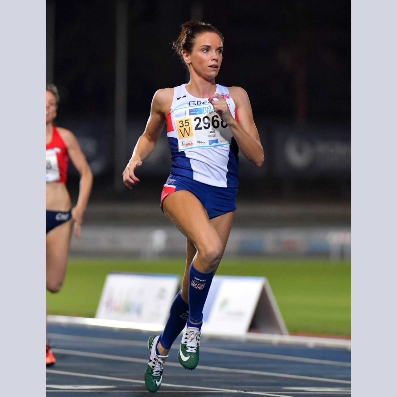 Lucy Evans running on an athletics tracks. Lucy is a white woman with dark hair. She wears blue shorts and white top. A competitor in red runs behind. The track is blue with a green field and crowd in the background.