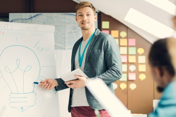A smiling white man dressed in business-casual clothing stands in front of flip chart, holding a digital tablet, while addressing colleagues sat around a table.