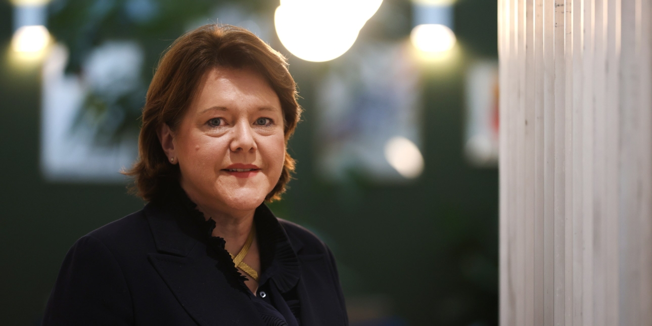 A colour portrait of Dame Maria Miller on against a blurred background with some lights behind. Maria Miller is a white woman with shoulder length brown hair