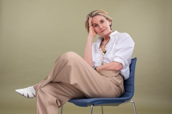 A colour photograph of Mary Portas sat in a chair against a beige background. Mary is a white woman with short blond hair wearing a white shirt and beige trousers.