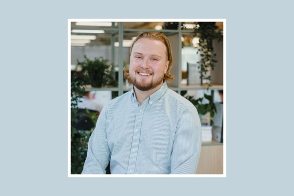 A white smiling man looks at the camera. He has ginger hair and a beard and is wearing a blue shirt and the background is an office