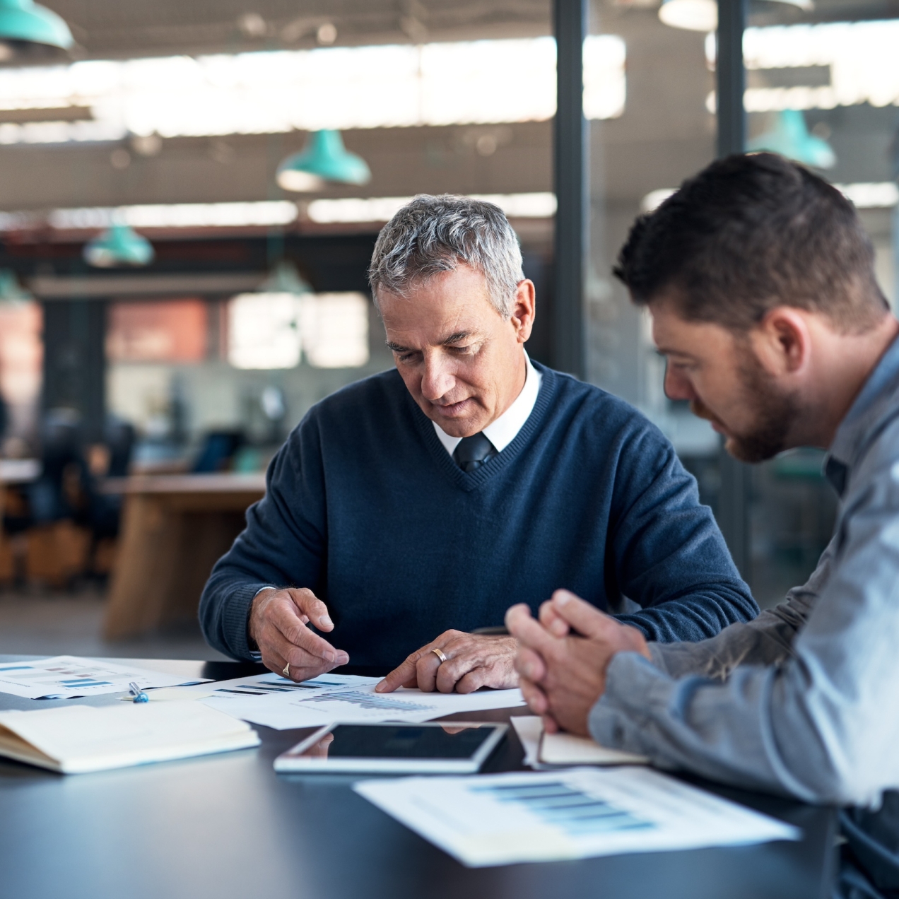 Two white men sit at a table analysing graphs. The man on the left is older with grey hair and a navy jumper. The man on the right has brown hair and beard and wears a light blue shirt.