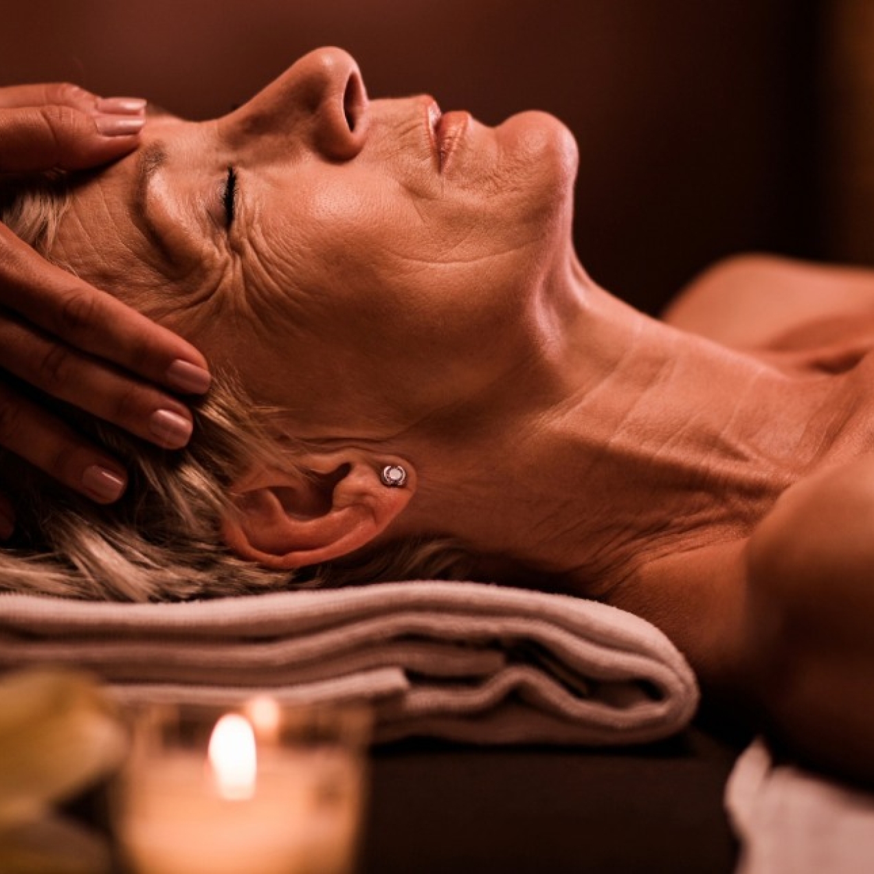 In a dark room lit by candles, a mature white woman with light coloured hair receives a head massage.