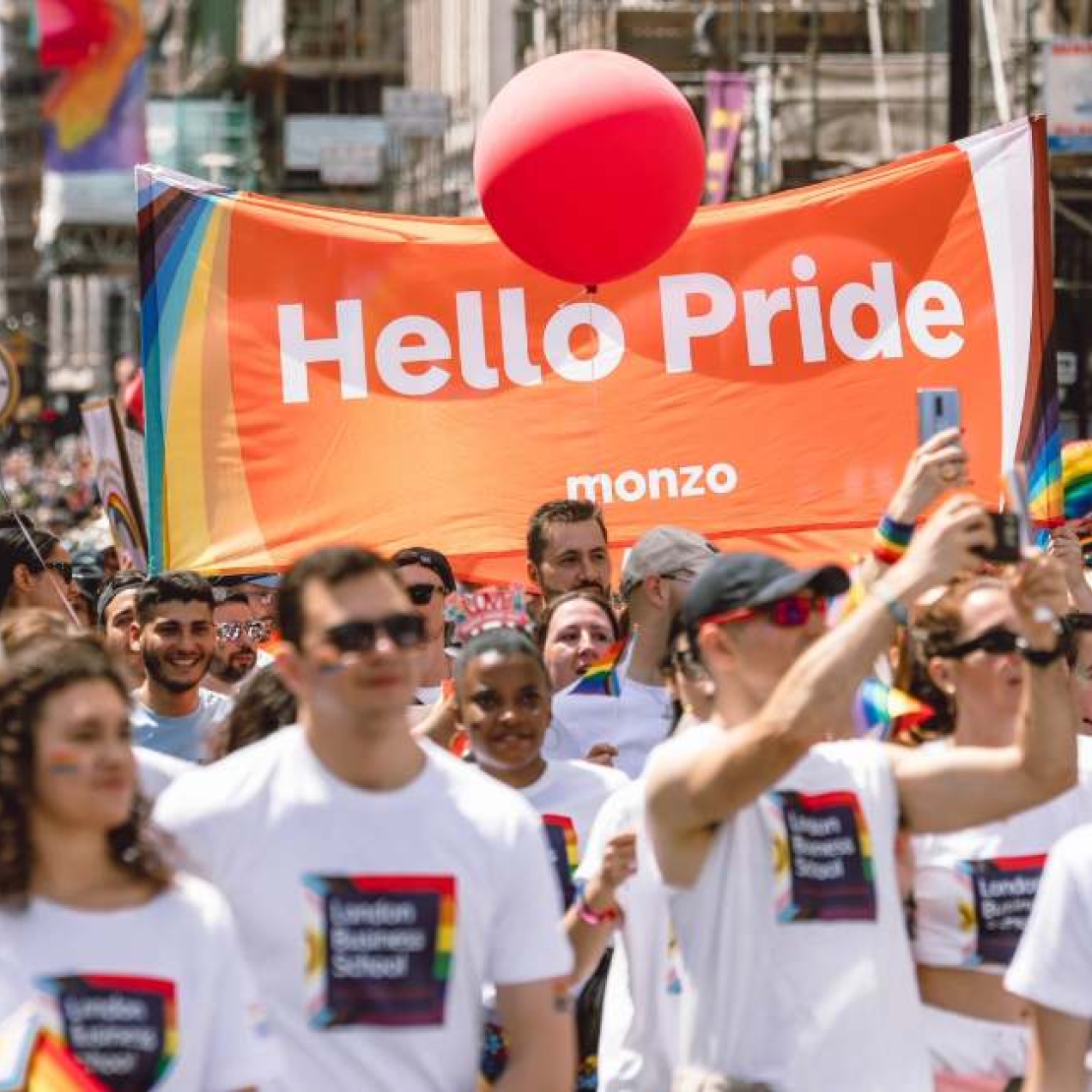 A group of people on a Pride march wearing white t-shirts. In the background an orange banner reads Hello Pride