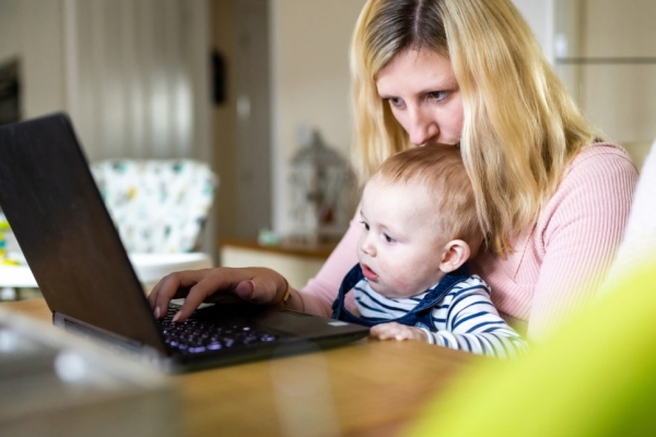 At a kitchen table a white woman with blonde hair kisses her baby's head sat in her lap while working at her laptop
