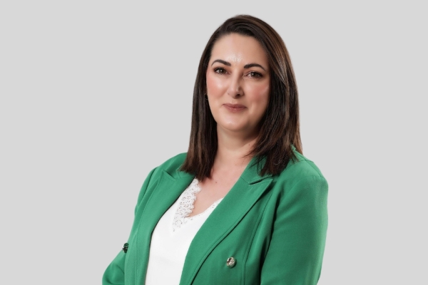 A colour portrait of Natasha Irvine against a grey background. Natasha is a white woman with shoulder length brown hair who wears a white top and green jacket. She smiles at the camera.