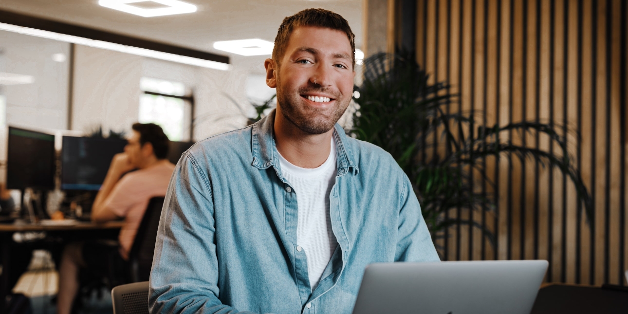 Ollie Manster, a white man with cropped brown hair, sat at a laptop smiling to camera. He wears a denim shirt over a white t-shirt, in modern office